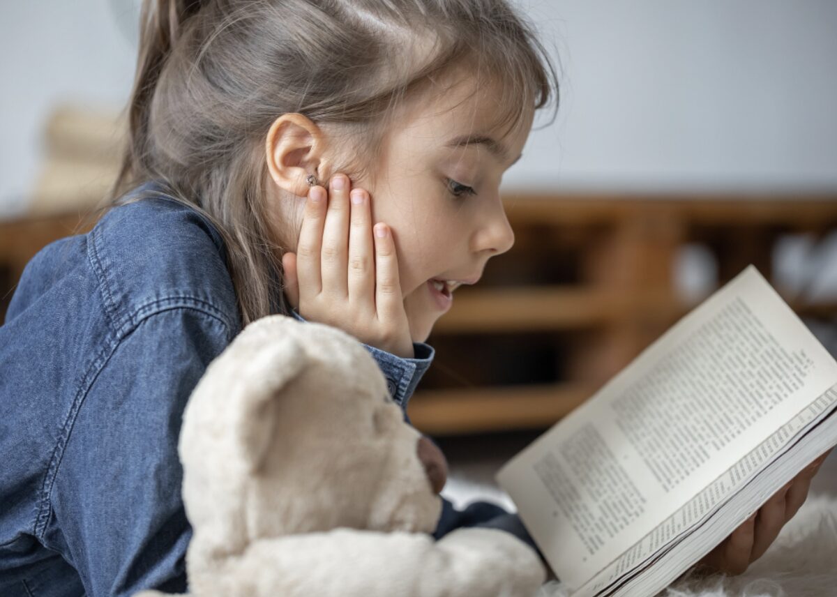 Jeune fille en train de lire un livre à voix haute avec sa peluche à côté d'elle