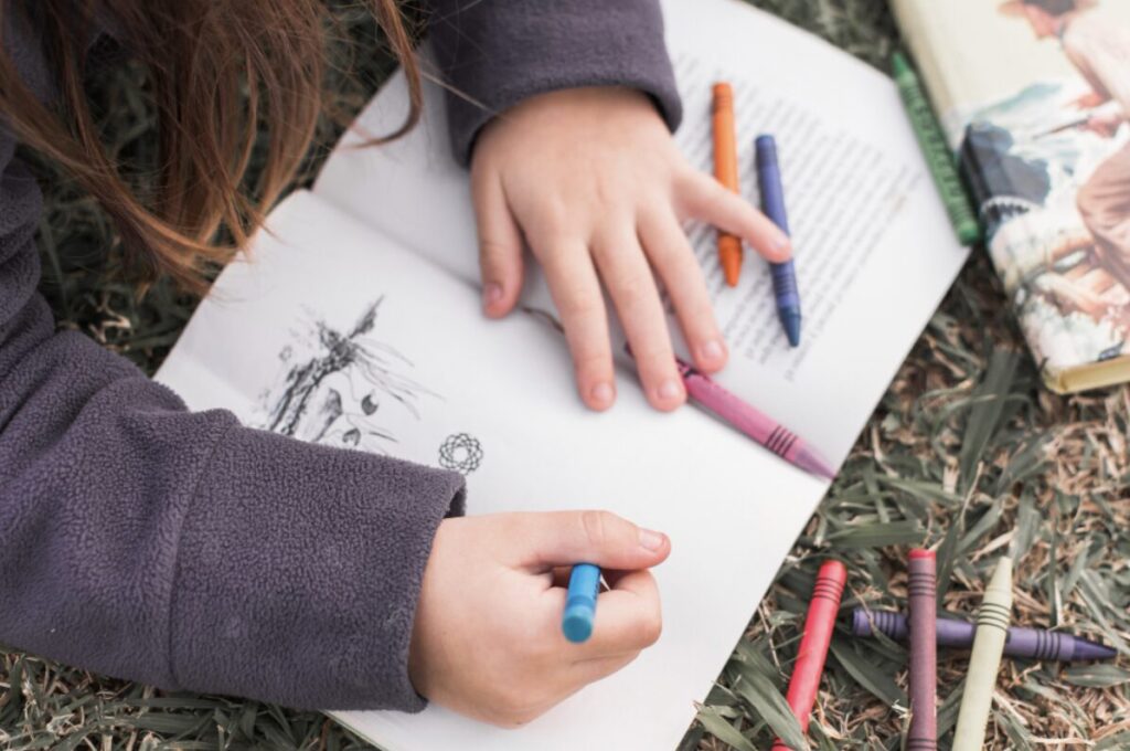 Mains d'un enfant en train de dessiner sur un cahier avec des crayons pastels de différentes couleurs