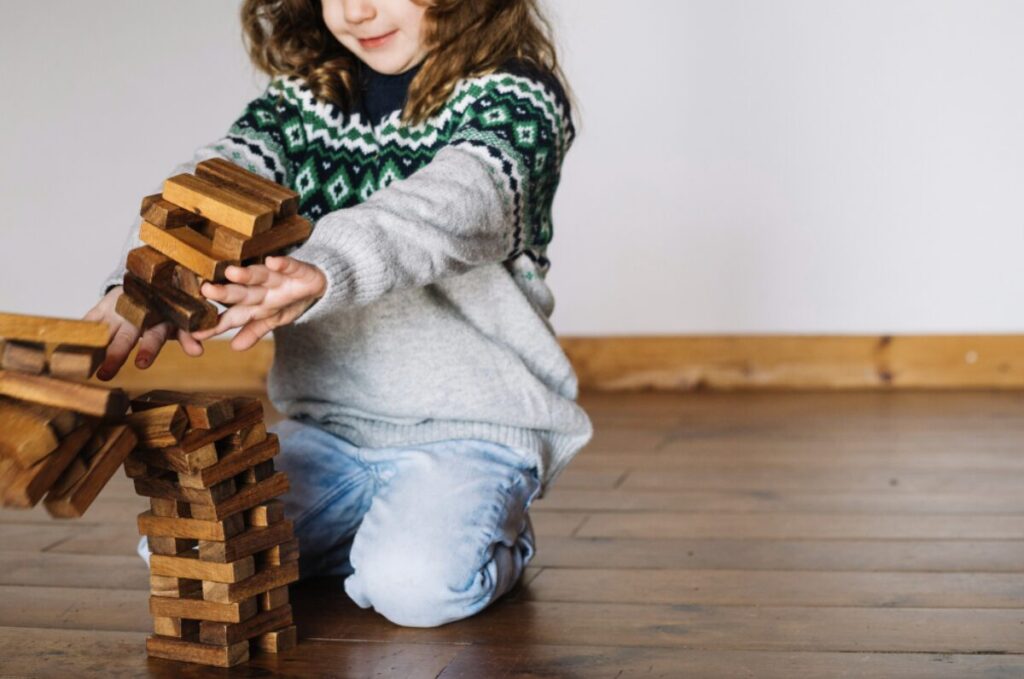 Jeune fille joyeuse, à genou dans une pièce avec un parqet au sol, en train de casser une tour faite avec des morceaux de bois