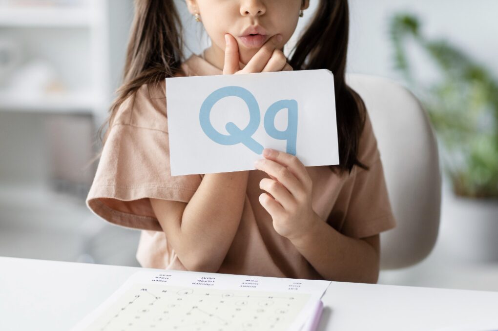 jeune fille tenant une carte dans sa main qu'elle montre pour qu'on la voie et sur laquelle se trouve écrites deux lettres Q, en majuscule et en minuscule