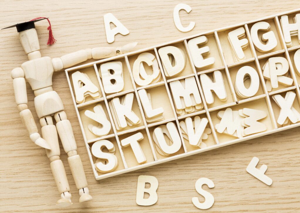 Boite de rangement contenant les lettres de l'alphabet et un bonhomme en bois articulé qui porte un chapeau de remise de diplôme et pose son bras sur la boîte